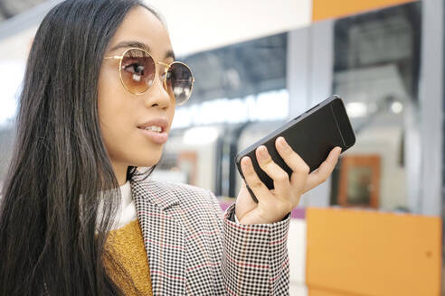 Nahaufnahme einer Frau mit Sonnenbrille, die am Bahnhof über ein Smartphone spricht - JCZF00128