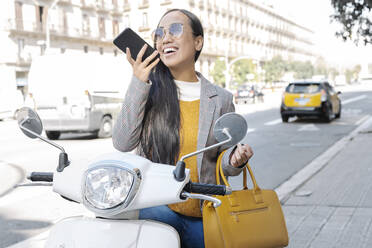 Fröhliche junge Frau, die über ihr Smartphone spricht, während sie auf einem Motorroller sitzt - JCZF00136