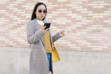 Stylish young woman wearing sunglasses using smart phone while standing against wall in city - JCZF00137