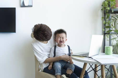 Mother holding crying boy on lap while sitting over chair at home - JPTF00509