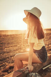 Thoughtful young woman sitting on retaining wall at field against clear sky during sunset - ACPF00745