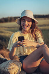 Fröhliche junge Frau, die während des Sonnenuntergangs auf einem Feld ihr Smartphone benutzt - ACPF00748