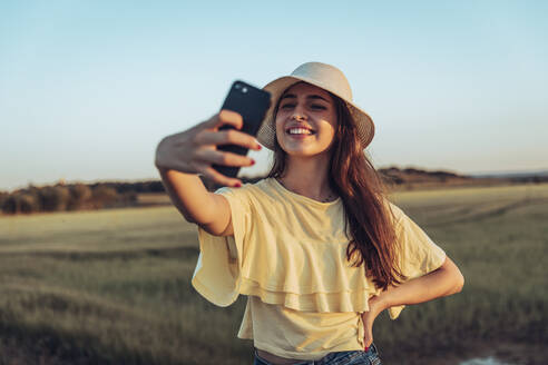 Glückliche junge Frau nimmt Selfie durch Smartphone gegen klaren Himmel bei Sonnenuntergang - ACPF00749