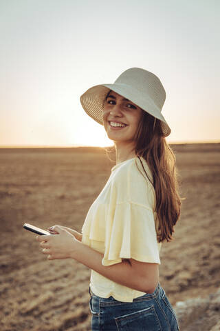 Glückliche junge Frau, die ihr Smartphone in der Hand hält, während sie auf einem Feld vor einem klaren Himmel bei Sonnenuntergang steht, lizenzfreies Stockfoto