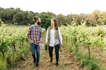 Happy couple holding wineglasses and wine bottle while walking at vineyard - JRFF04487