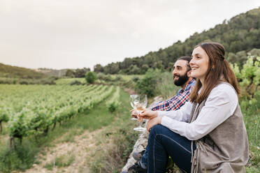 Happy couple holding white wine glasses while sitting at vineyard - JRFF04495