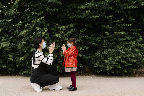 Mother and daughter wearing masks while playing clapping game on street - EGAF00123