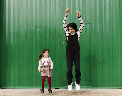 Playful mother and daughter jumping against green corrugated wall - EGAF00132