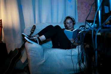 Female musician reading book while sitting on sofa in recording studio at home - ERRF03958