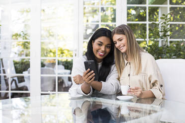 Glückliche multiethnische Freundinnen machen ein Selfie mit ihrem Smartphone am Tisch in einem Café - ABZF03167