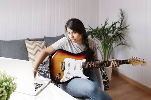 Junge Frau benutzt einen Laptop, während sie zu Hause E-Gitarre spielt - JMHMF00051