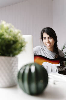 Happy young woman using laptop while sitting with electric guitar at home - JMHMF00052