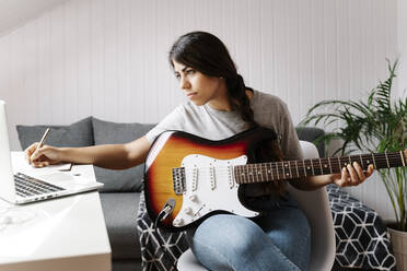 Young woman writing while using laptop and electric guitar at home - JMHMF00055