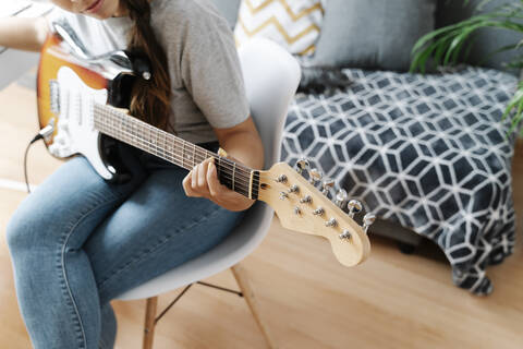 Frau spielt E-Gitarre, während sie auf einem Stuhl zu Hause sitzt, lizenzfreies Stockfoto