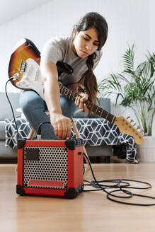 Woman adjusting knobs on amplifier while playing electric guitar at home - JMHMF00058