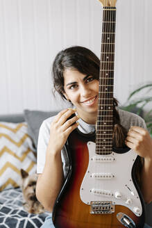 Happy woman holding electric guitar while sitting on sofa at home - JMHMF00059