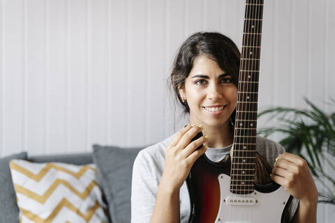 Confident young woman holding electric guitar while sitting on sofa at home stock photo