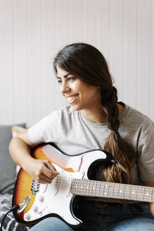 Happy woman playing electric guitar while sitting at home - JMHMF00061