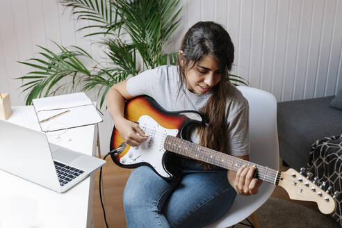 Young woman playing electric guitar while sitting on chair at home - JMHMF00062