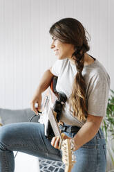 Happy woman playing electric guitar while standing at home - JMHMF00063