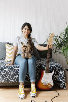 Happy young woman holding electric guitar while sitting with Yorkshire Terrier on sofa at home - JMHMF00064
