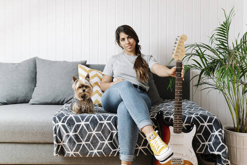 Confident woman holding electric guitar while sitting with Yorkshire Terrier on sofa at home - JMHMF00065