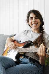 Fröhliche Frau spielt E-Gitarre, während sie zu Hause sitzt - JMHMF00066