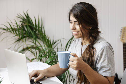 Glückliche junge Frau, die einen Kaffeebecher hält, während sie zu Hause einen Laptop benutzt, lizenzfreies Stockfoto