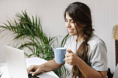 Happy young woman holding coffee mug while using laptop at home - JMHMF00068