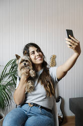 Junge Frau macht Selfie mit Yorkshire Terrier, während sie zu Hause auf einem Stuhl sitzt - JMHMF00069