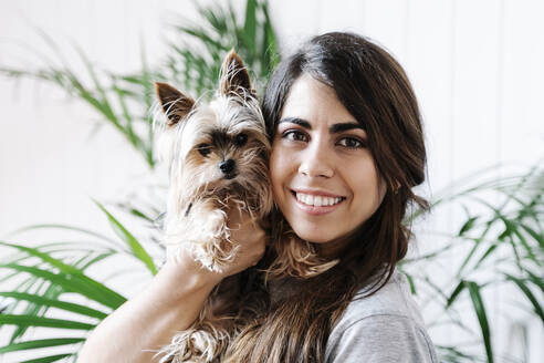 Happy young woman carrying Yorkshire Terrier at home - JMHMF00070