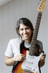 Glückliche Frau hält E-Gitarre gegen die Wand zu Hause - JMHMF00074