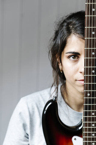 Junge Frau versteckt sich hinter einer E-Gitarre an der Wand zu Hause, lizenzfreies Stockfoto