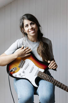 Happy young woman holding electric guitar while sitting at home - JMHMF00077