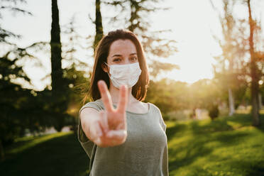 Portrait of woman wearing protective mask in nature showing victory sign - EBBF00181