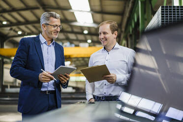Zwei lächelnde Geschäftsleute mit Tablet und Klemmbrett in einer Fabrik - MOEF02915
