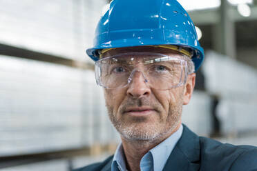 Porträt eines reifen Geschäftsmannes mit Schutzhelm und Schutzbrille in einer Fabrik - MOEF02948