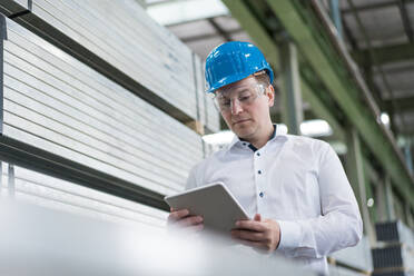 Businessman wearing hard hat and safety goggles in a factory using tablet - MOEF02950