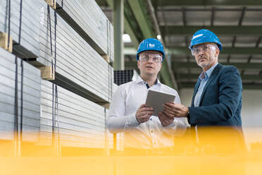 Two businessmen wearing hard hats and safety goggles in a factory using tablet - MOEF02952