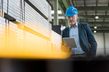 Mature businessman wearing hard hat and safety goggles in a factory using tablet - MOEF02953