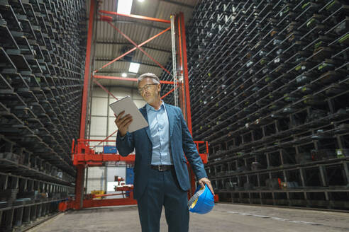 Mature businessman using tablet in a high rack warehouse of a factory - MOEF02970