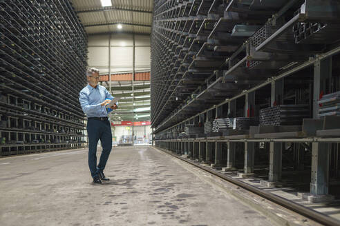 Mature businessman using tablet in a high rack warehouse of a factory - MOEF02977