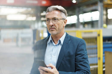 Mature businessman holding mobile phone in a factory - MOEF03012