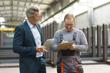 Happy businessman with tablet and employee with clipboard in a factory - MOEF03053