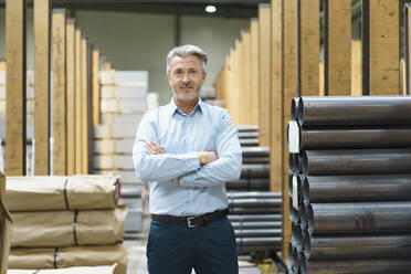 Portrait of a confident mature businessman in a factory storehouse with steel pipes - MOEF03076