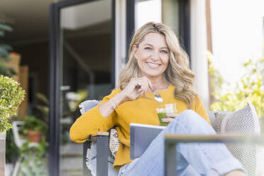 Porträt einer glücklichen reifen Frau, die mit einem digitalen Tablet auf der Terrasse sitzt und einen grünen Smoothie genießt - UUF20531