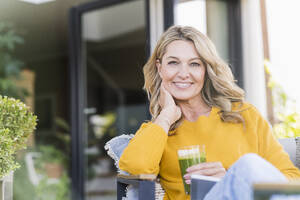 Porträt einer glücklichen reifen Frau, die mit einem digitalen Tablet und einem grünen Smoothie auf der Terrasse sitzt - UUF20532