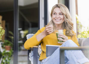 Porträt einer lächelnden reifen Frau, die mit einem digitalen Tablet auf einer Terrasse sitzt und einen grünen Smoothie genießt - UUF20534