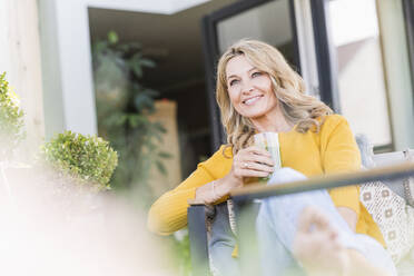 Porträt einer lächelnden reifen Frau, die mit einem grünen Smoothie auf der Terrasse sitzt - UUF20535