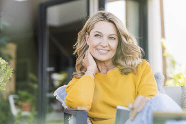 Porträt einer lächelnden reifen Frau, die mit einem digitalen Tablet auf einer Terrasse sitzt und in die Ferne schaut - UUF20539